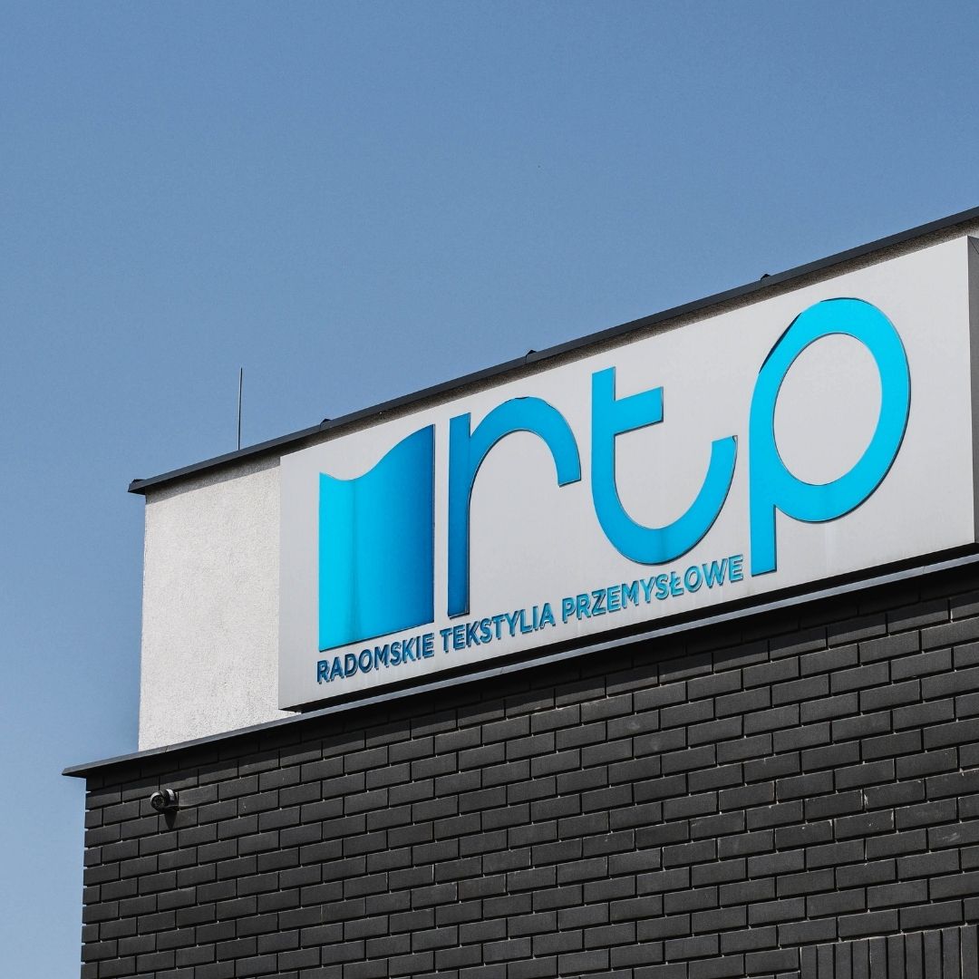 Logotyp firmy RTP Radom, czyli Radomskie Tekstylia Przemysłowe, umieszczony na elewacji nowoczesnego budynku przemysłowego.